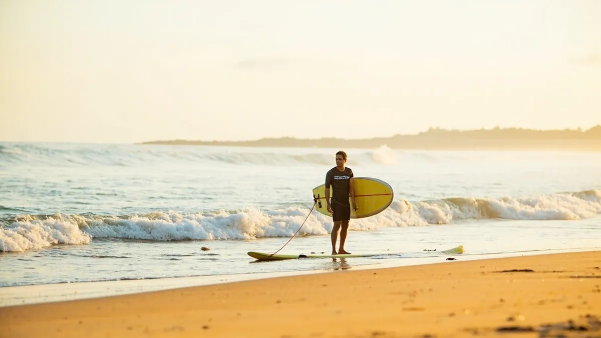 Beginner surfer learning on a foam board in gentle waves at Serenity Beach with an instructor beside them.